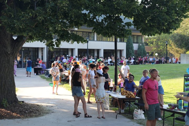 Students and community members gather at an outdoor activities fair, with tables set up along a path and people chatting, browsing, and interacting with various booths. The event takes place on a sunny day near a large campus building.